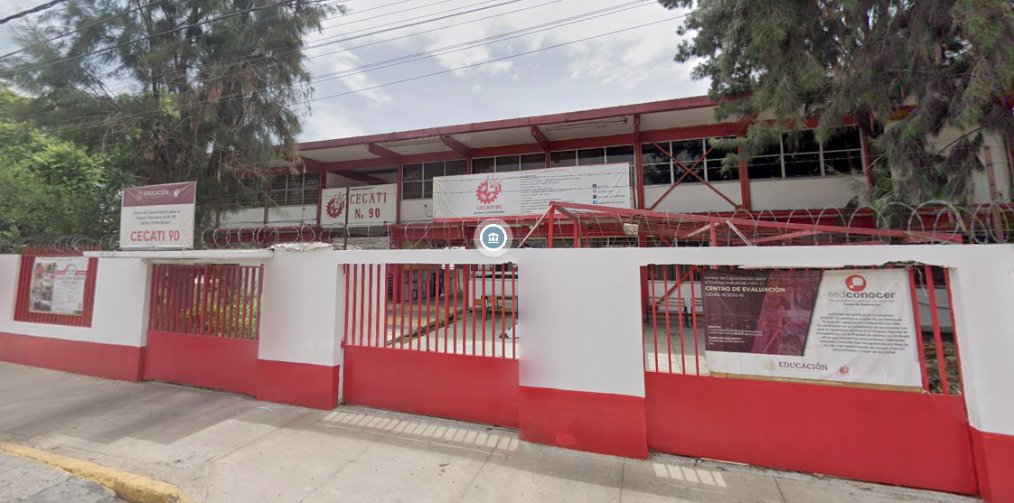 Fotografía en la que se muestra la entrada del plantel, conformada por muros blancos intercalados con rejas rojas. Encima de una de las rejas hay una placa que dice en letras rojas CECATI 90.